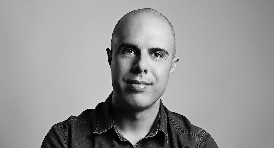 Um homem careca com uma camisa jeans está sorrindo em uma foto em preto e branco.
