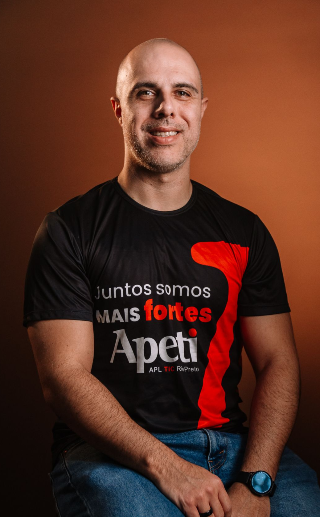 Um homem careca está sentado vestindo uma camisa preta e laranja.