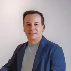 Um homem de jaqueta azul e camisa cinza está sorrindo para a câmera.