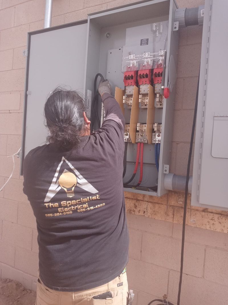 A person in a black shirt working on the internal wiring of an open electrical service panel mounted on a brick wall.