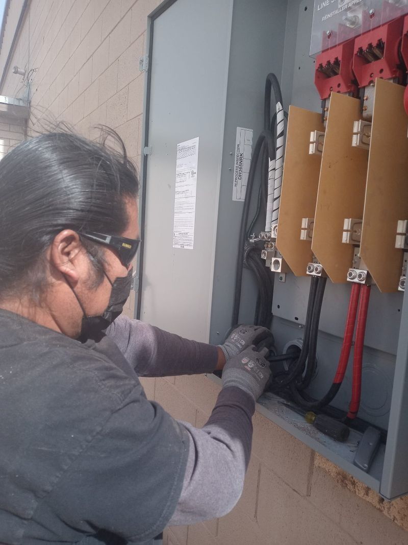 A technician wearing protective eyewear and gloves works on the wiring inside an open electrical panel.