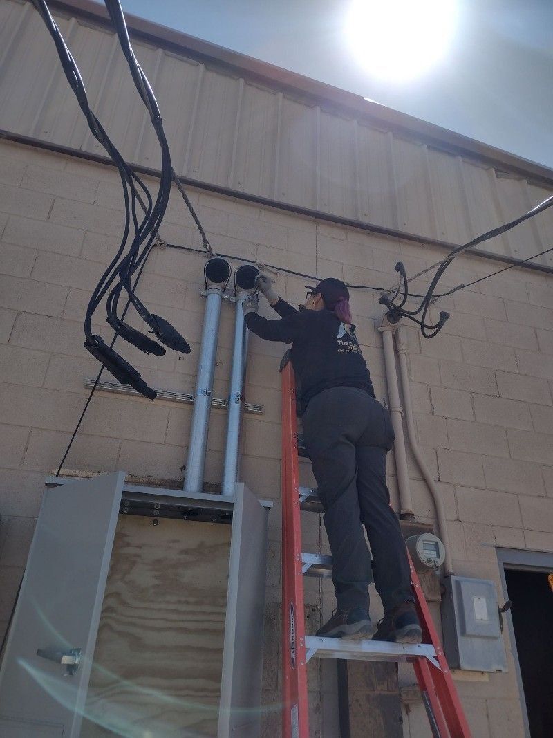 An individual in dark clothing stands on a ladder working on electrical conduits mounted to a beige concrete wall.