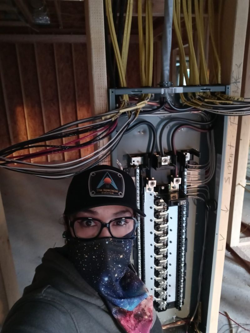 A person wearing a patterned face covering and baseball cap takes a selfie in front of an unfinished electrical panel.