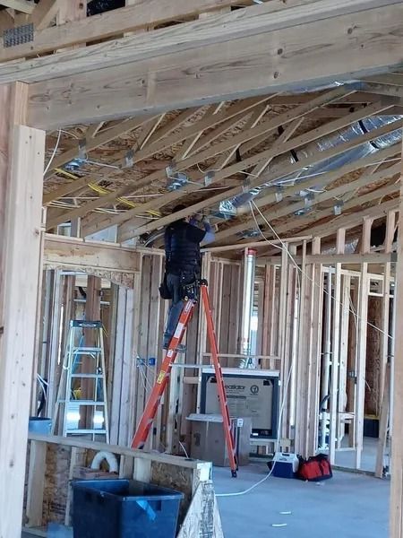 A worker stands on an orange stepladder, installing electrical wiring in the exposed wooden frame