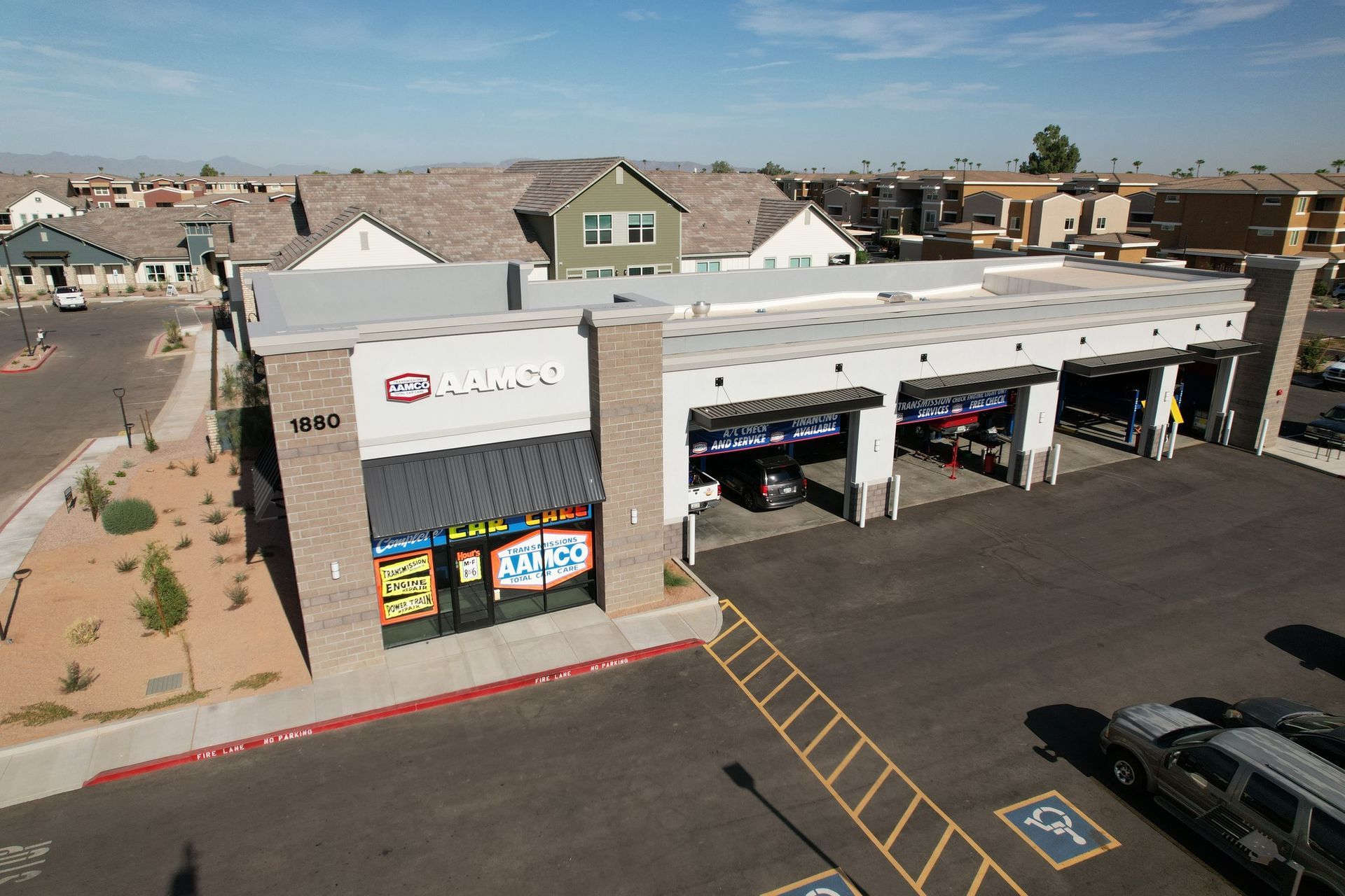 Exterior of an AAMCO Transmissions & Total Car Care shop with open bays, next to a parking lot and residential homes.