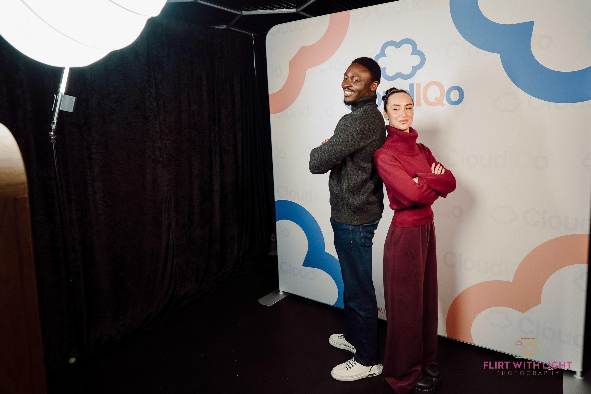 Two people pose back-to-back in front of a white and blue branded event backdrop.