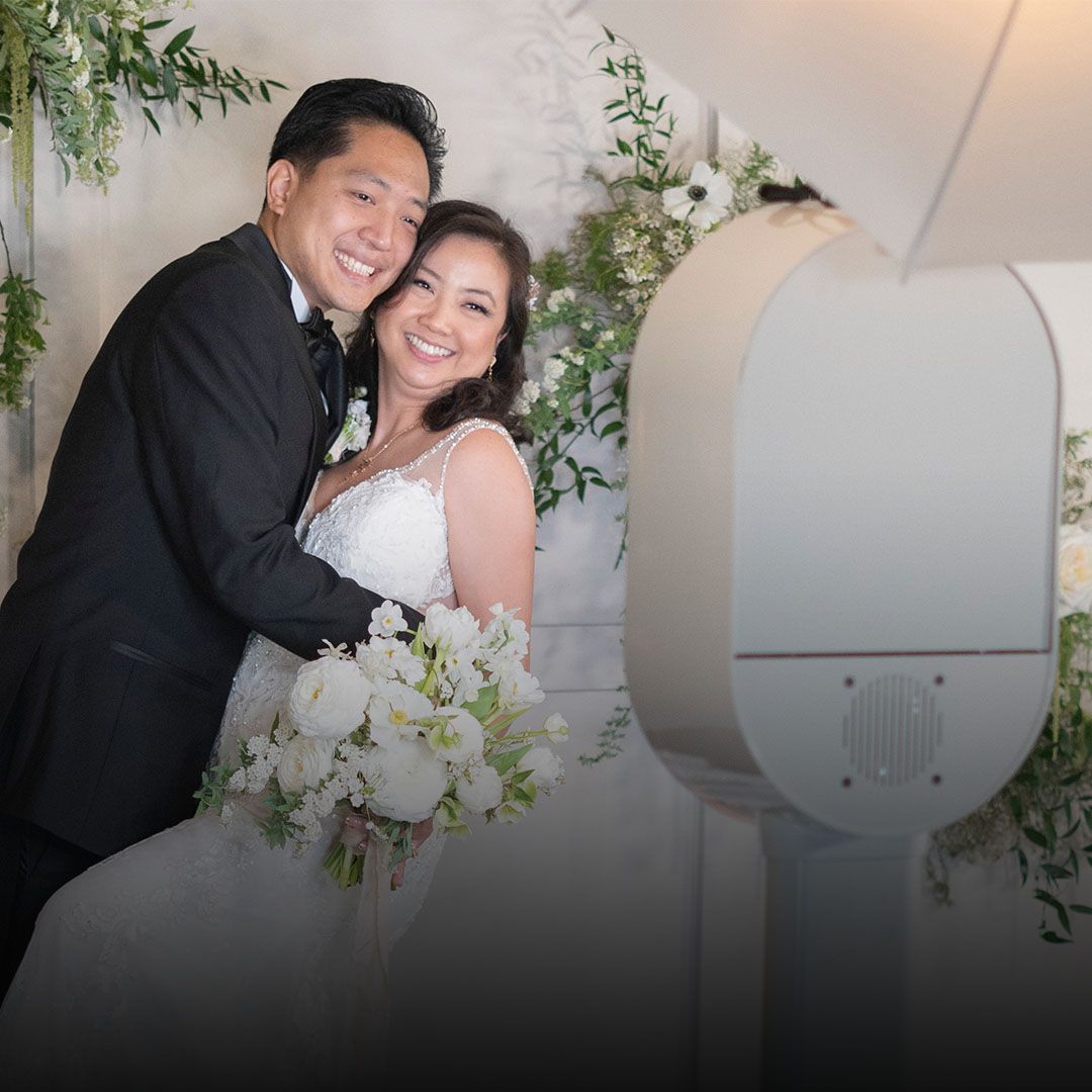 Smiling couple in wedding attire posing with a bouquet beside a large photo booth kiosk