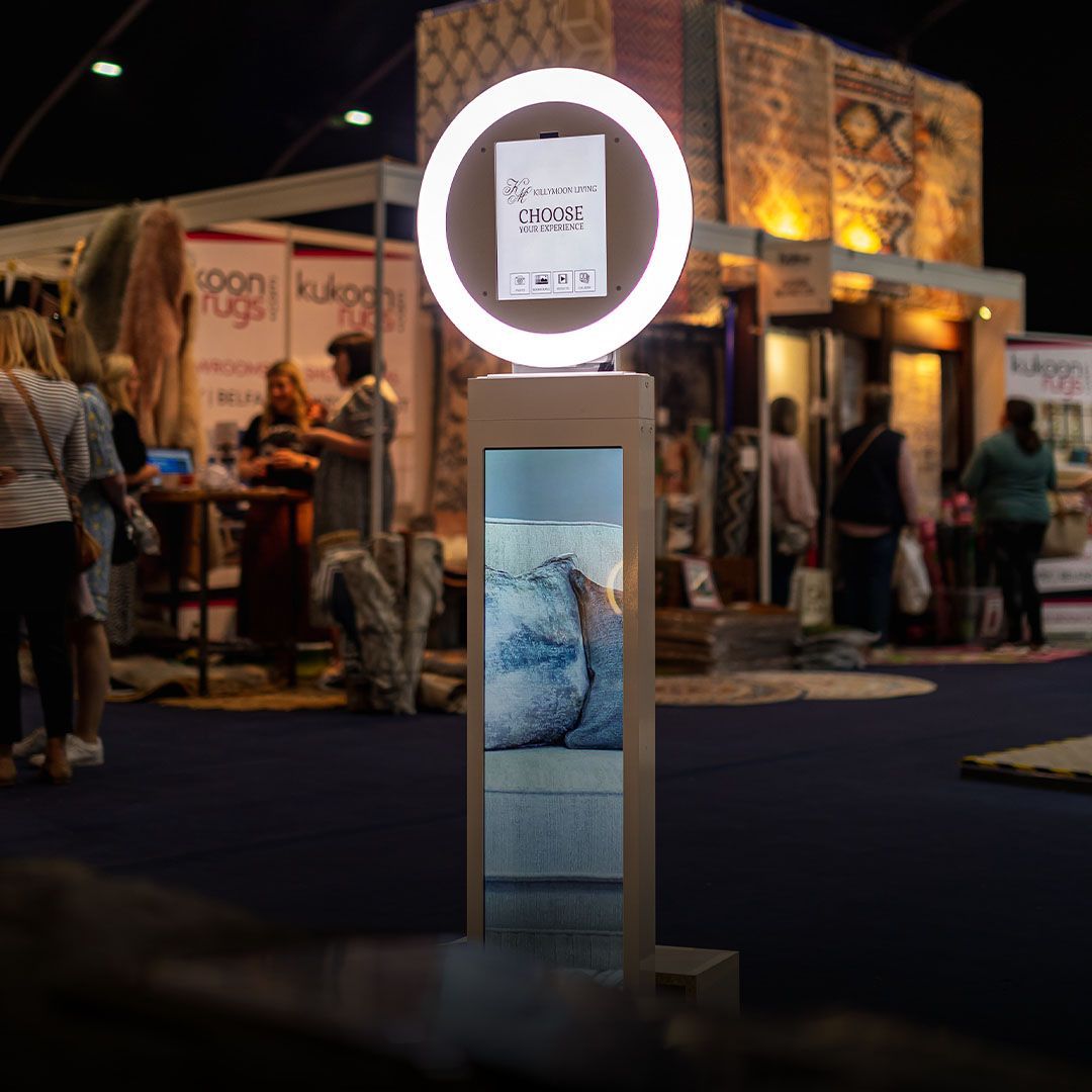 Circular digital kiosk with a lit ring screen at an indoor trade show booth, people blurred in background