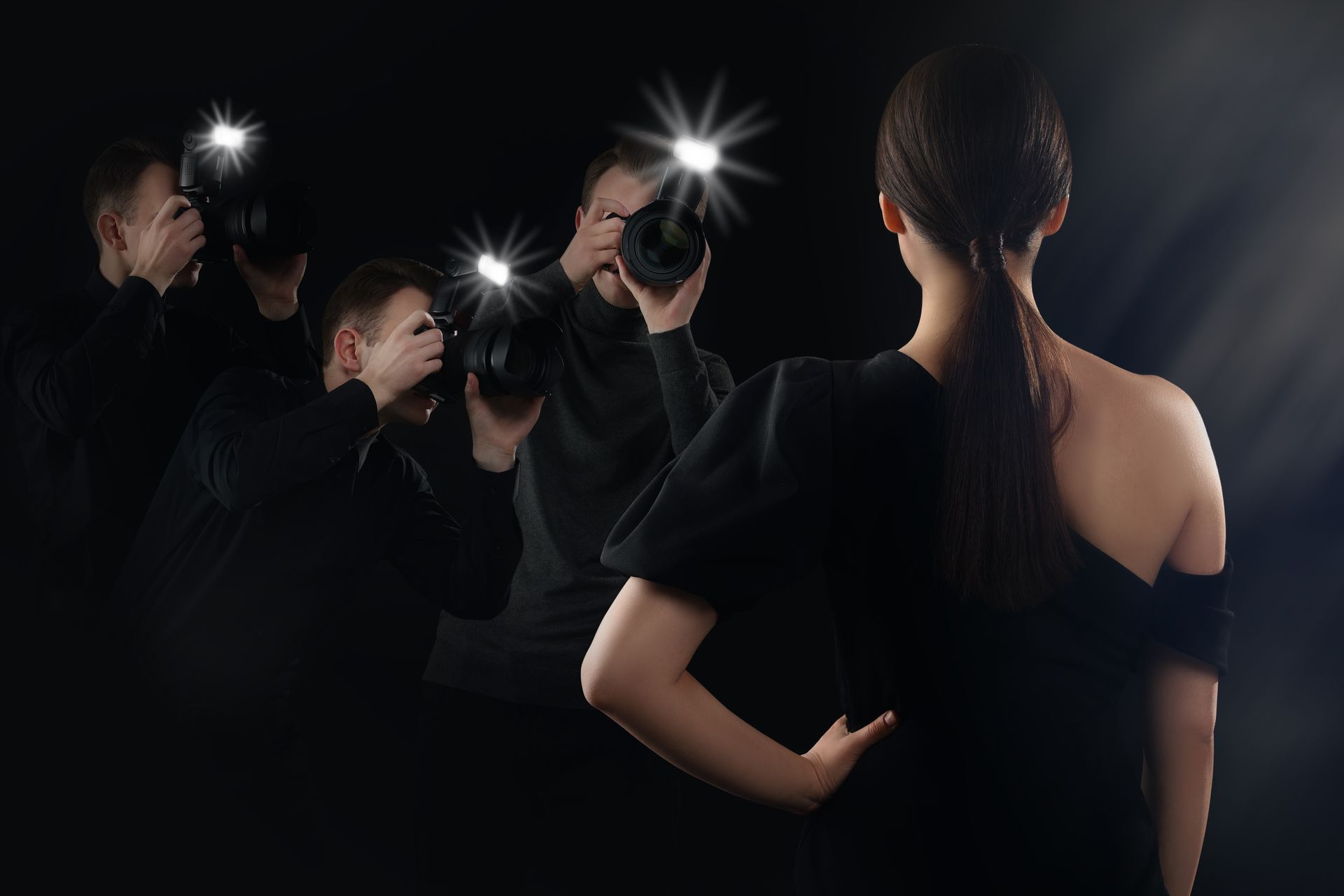 Photographers with flash cameras capturing a woman in a black dress, seen from behind against a dark background