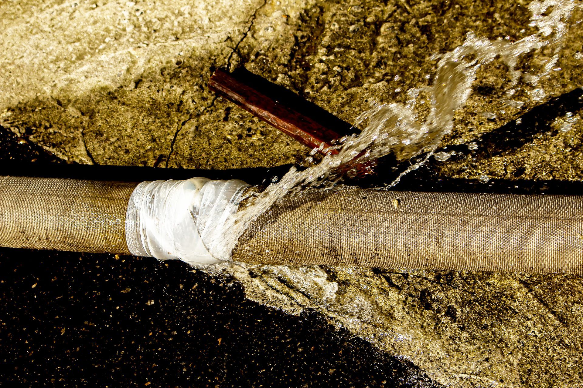 Water spraying from a leak in a mesh-covered hose that has been partially repaired with white tape.