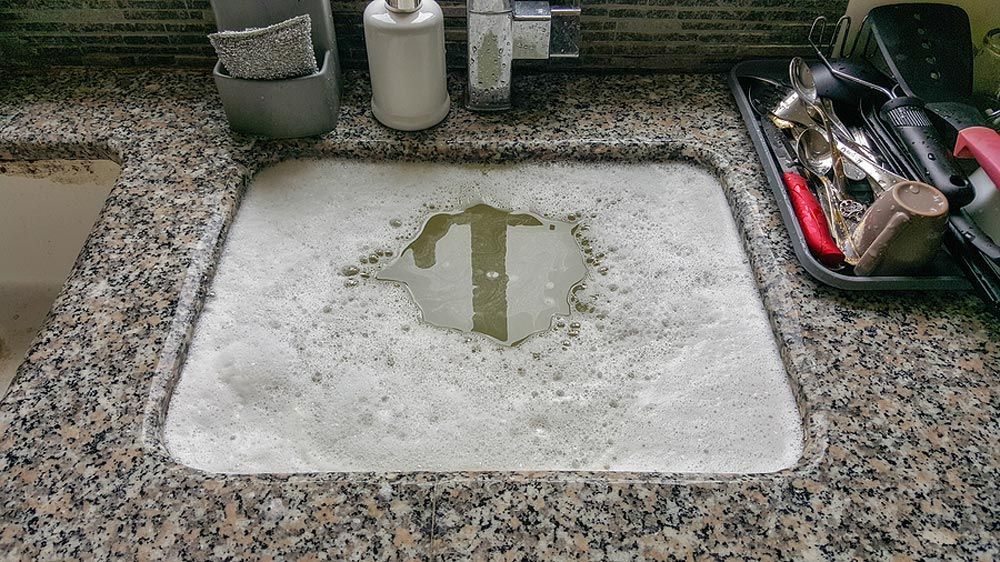 A kitchen sink filled with white soapy bubbles and a clear patch of water, with nearby bottles and cleaning supplies.