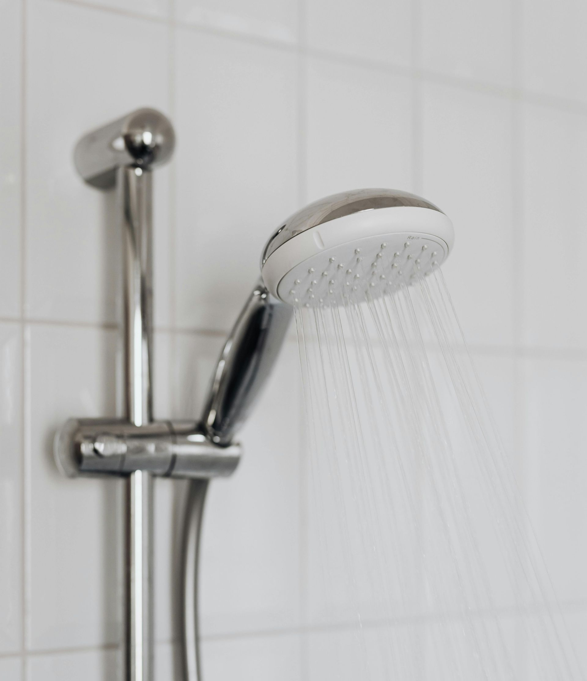 A chrome handheld showerhead attached to a vertical sliding bar, spraying water against a background of white tiled walls.