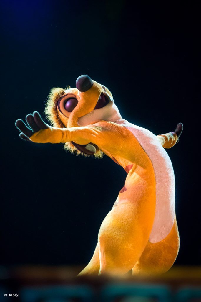 Animated meerkat-like animal singing on stage, lit by warm spotlight against a dark background