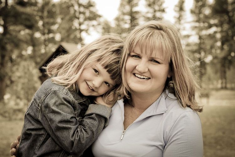 Smiling woman holding a young child outdoors in a wooded area.