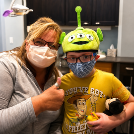 Masked adult and child indoors, smiling and giving thumbs up; child wears a green alien hat.