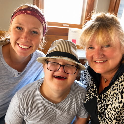 Three smiling people pose together indoors by a bright window.