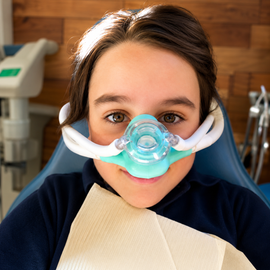 Person in a dental chair wearing a blue breathing mask, holding a bib, in a clinic office