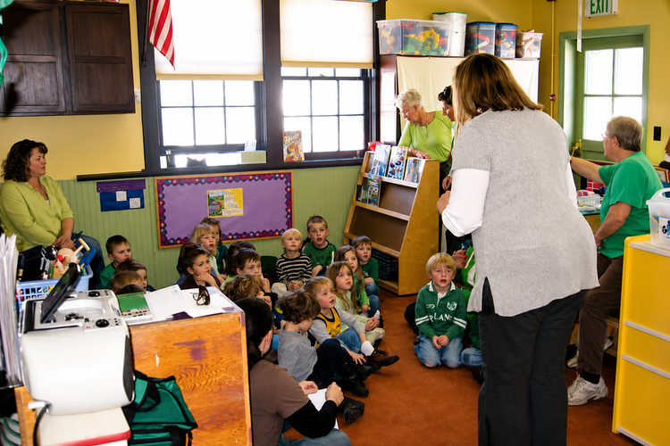 Teacher addresses seated children in a colorful classroom.