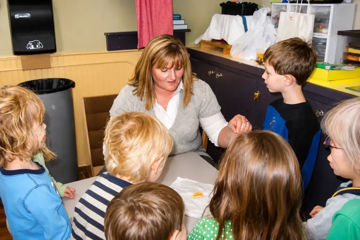 Teacher sitting with children in a classroom, speaking with them around a table.