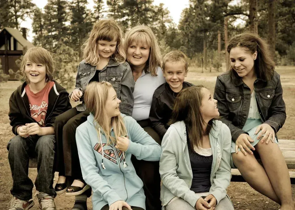 Seven friends sit and smile outdoors in a wooded area, with one leaning back and others looking at each other.