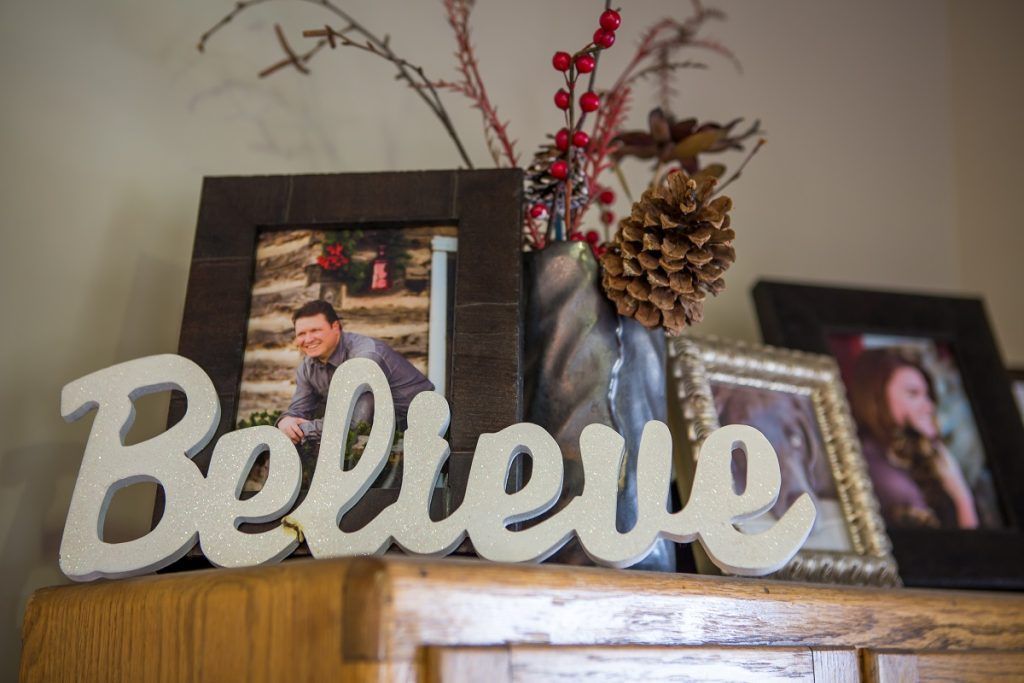 Shelf display with white “Believe” sign, framed photos, and pinecones in a vase with red berries.