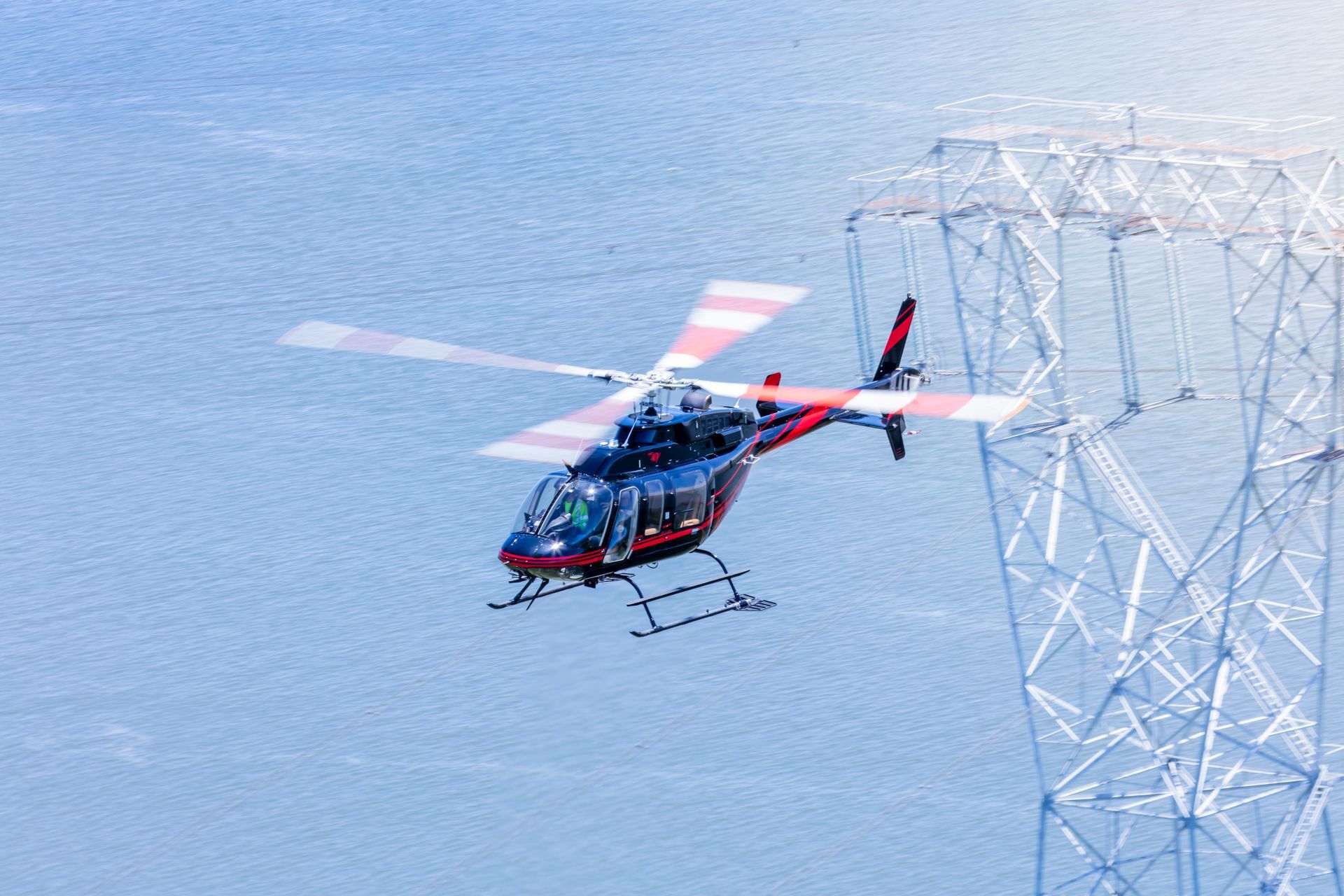 A helicopter is flying over a body of water near a power tower.