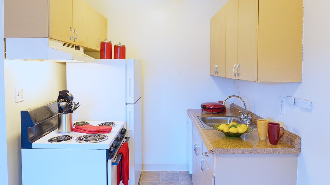Small kitchen with white stove, fridge, yellow cabinets, and a countertop with fruit and dishes.