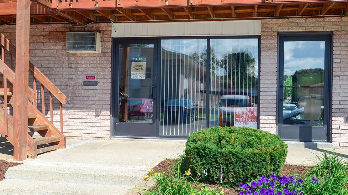 Storefront with glass doors, brick facade, steps, and a small landscaped bush in front.