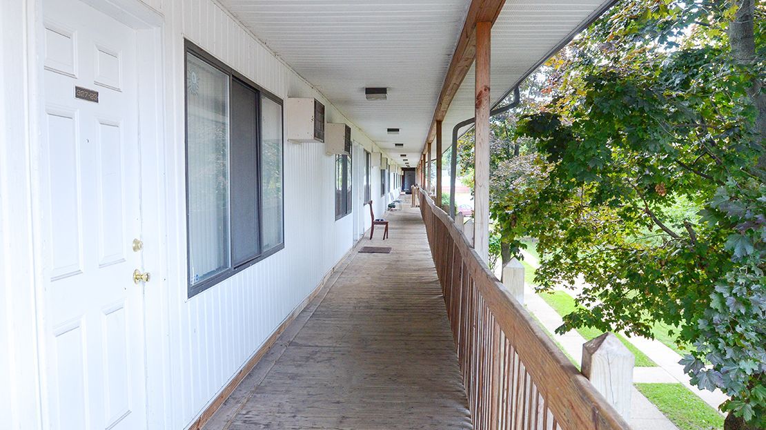 Covered apartment walkway with white doors, windows, and wooden railing beside trees and greenery.