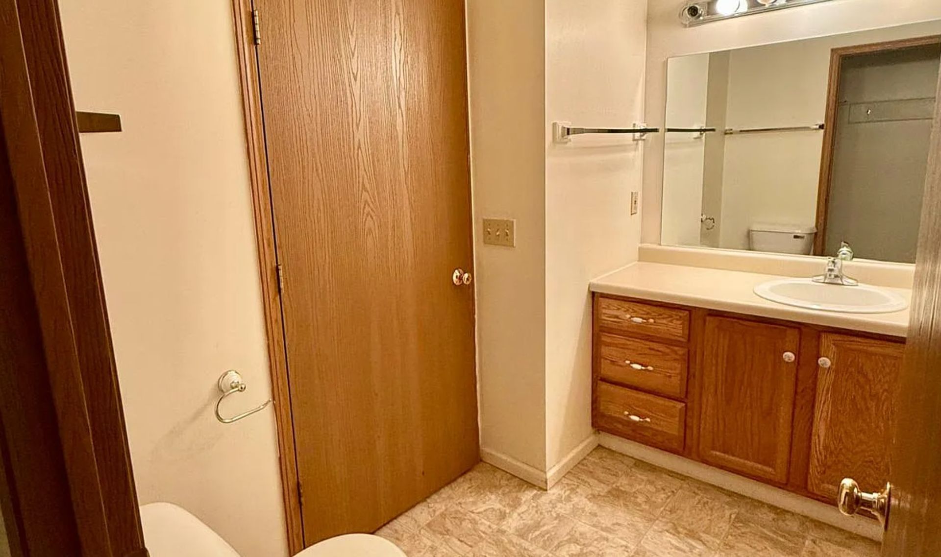 Bathroom with wood door, white sink vanity, mirror, and beige tile floor.