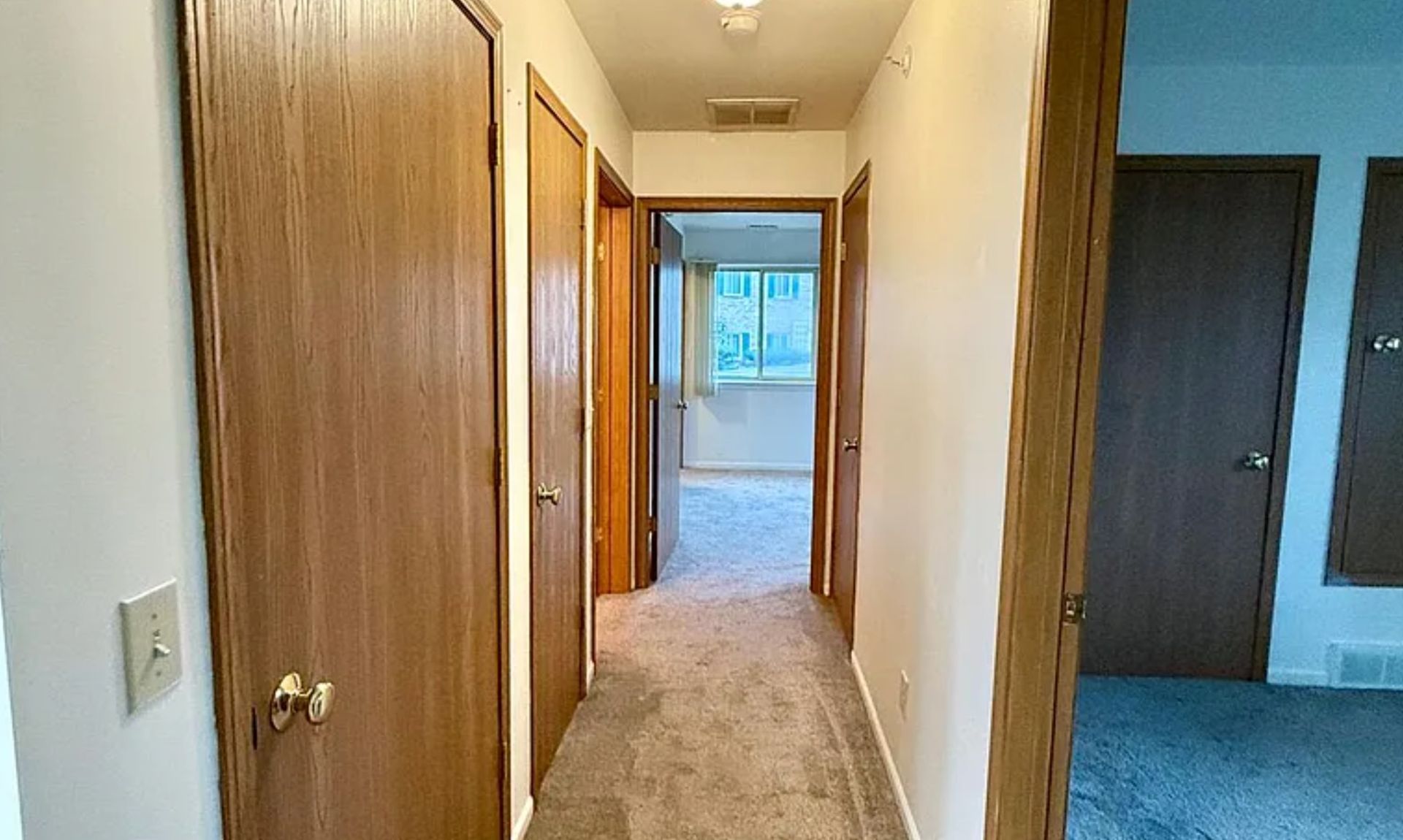 Narrow hallway with wood doors and beige carpet, leading to a bright room at the end.