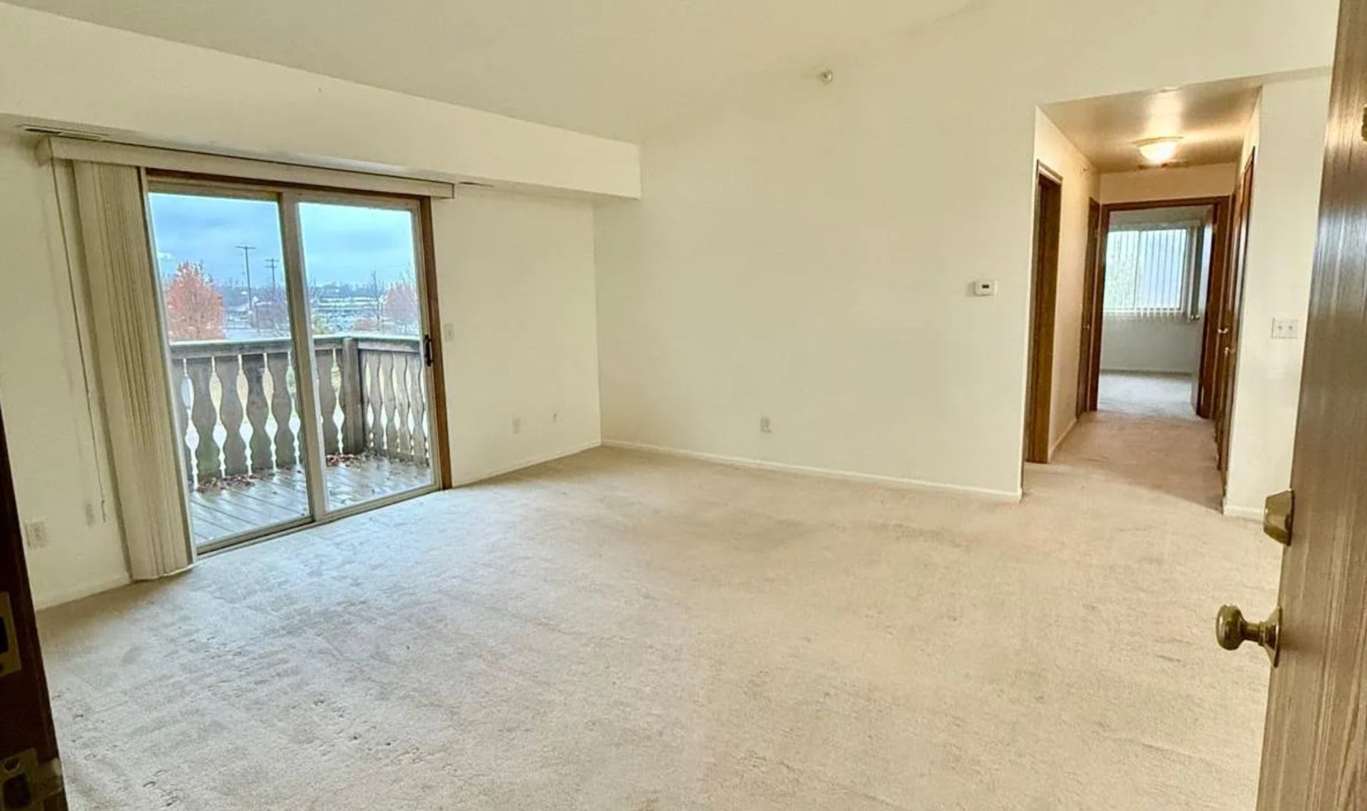 Empty beige living room with carpet, sliding glass doors to a balcony, and a hallway opening on the right