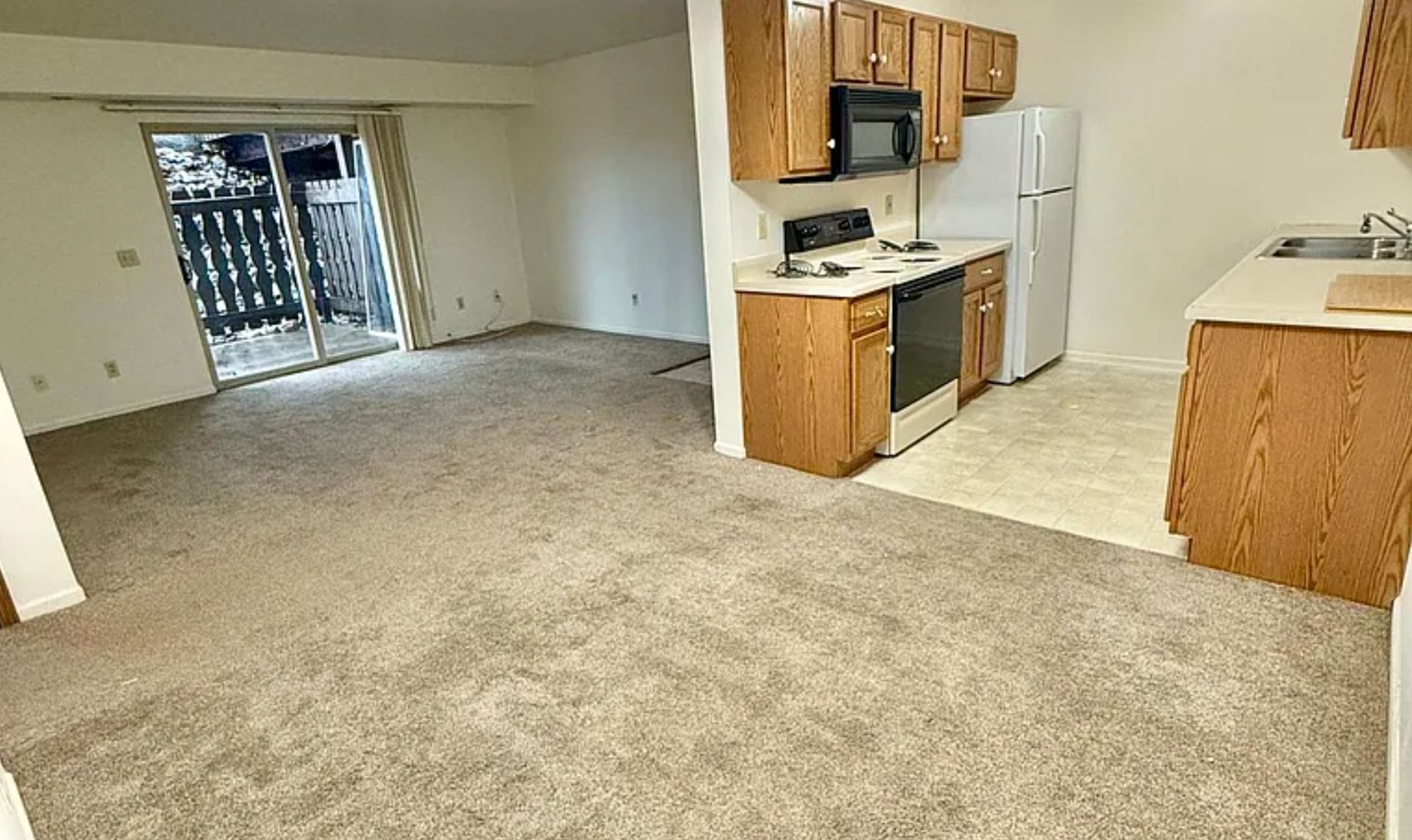 Empty apartment living area with beige carpet and open kitchen, white appliances, and sliding glass door to balcony