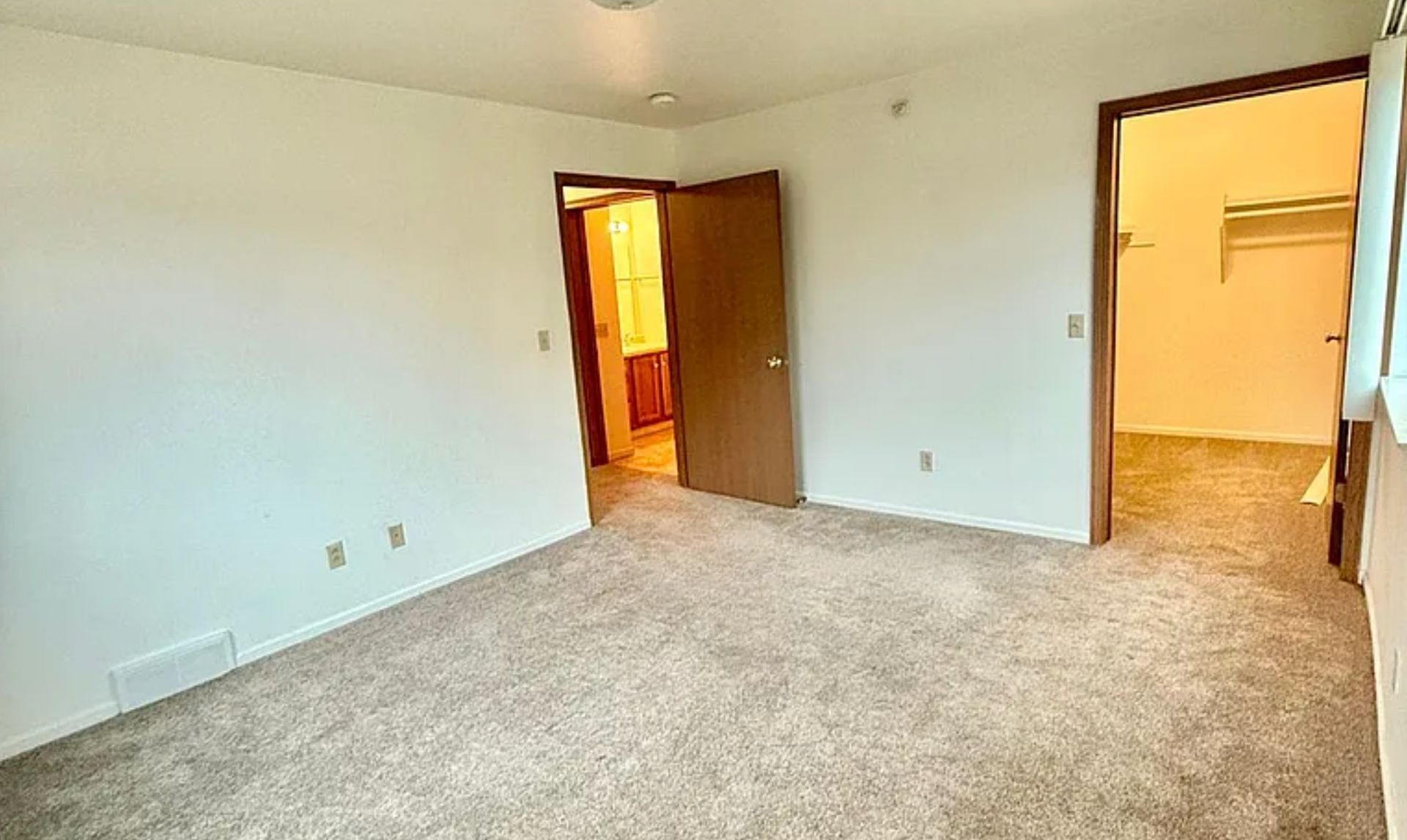 Empty carpeted bedroom with white walls, two open doors, and warm light from adjacent rooms