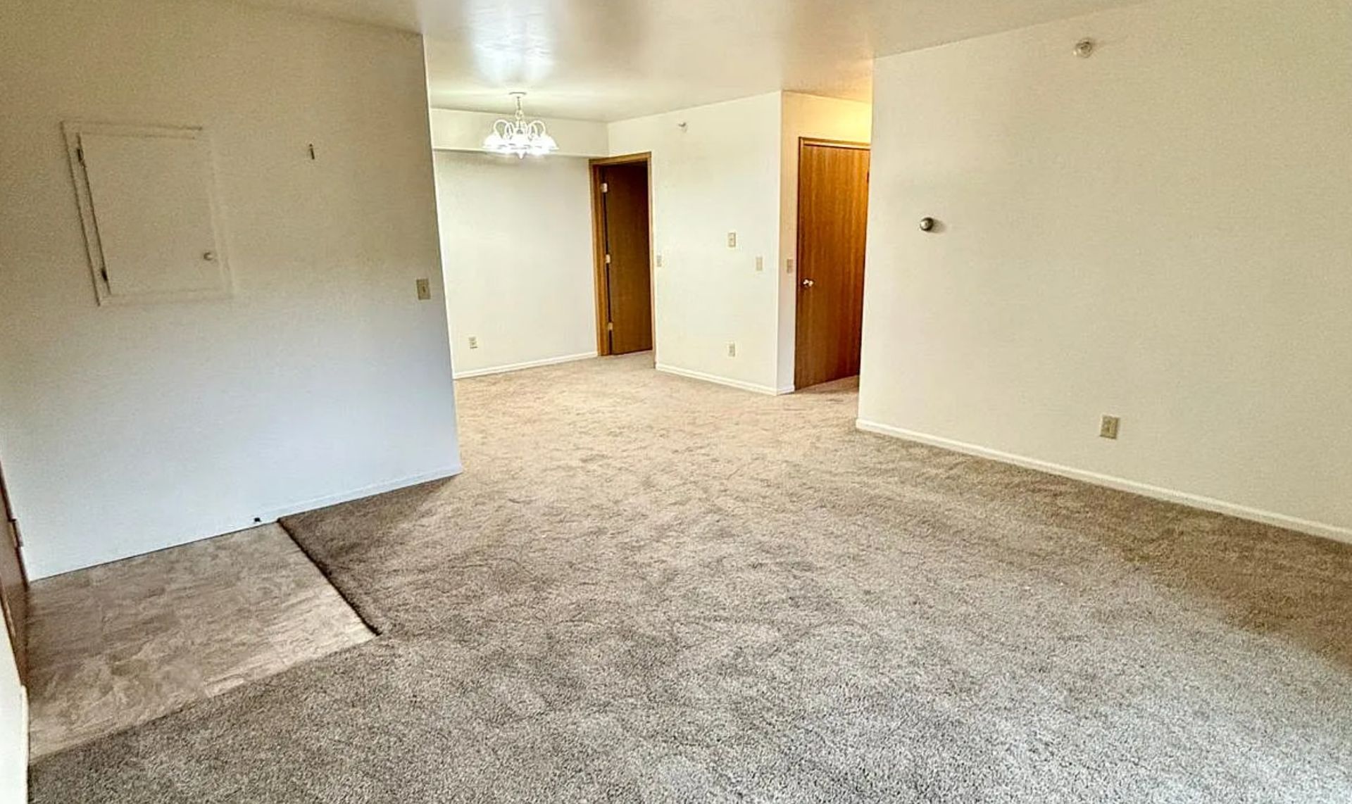 Empty carpeted living room with beige walls, white ceiling, and two wooden doorways.