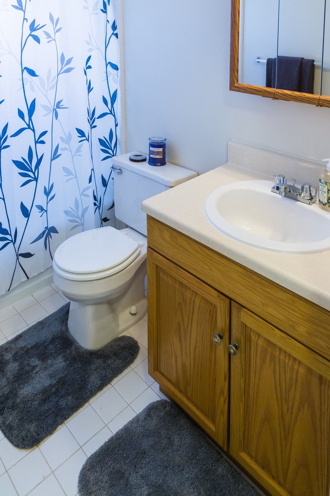 Small bathroom with white toilet, wood vanity, white sink, blue floral shower curtain, and gray rugs.