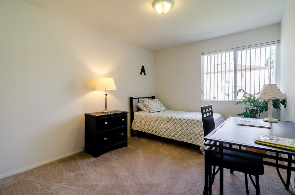 Small bedroom with twin bed, desk, nightstand, lamp, and window blinds