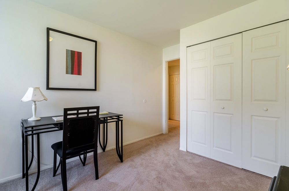 Small home office with black desk and chair, lamp, framed art, and white closet doors on carpet.