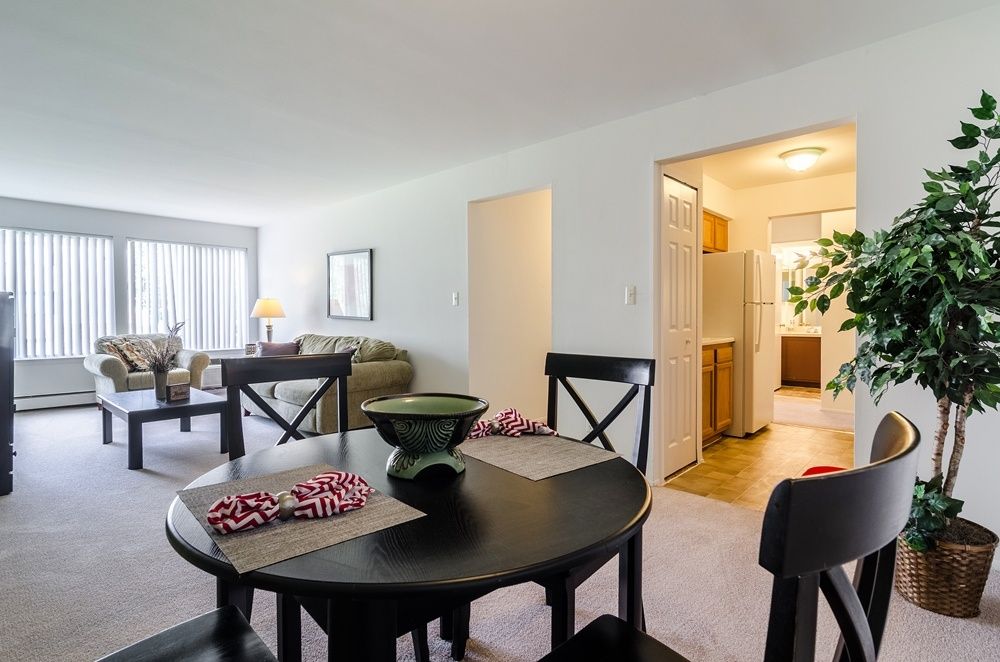 Bright open living and dining room with black tables, gray sofas, and a view into a lit kitchen.