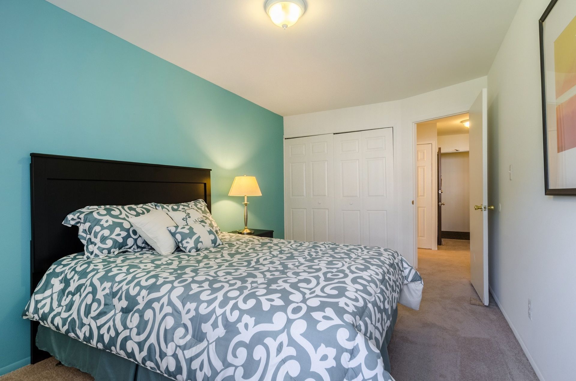 Bedroom with teal accent wall, patterned bedspread, and lamp beside an open doorway.