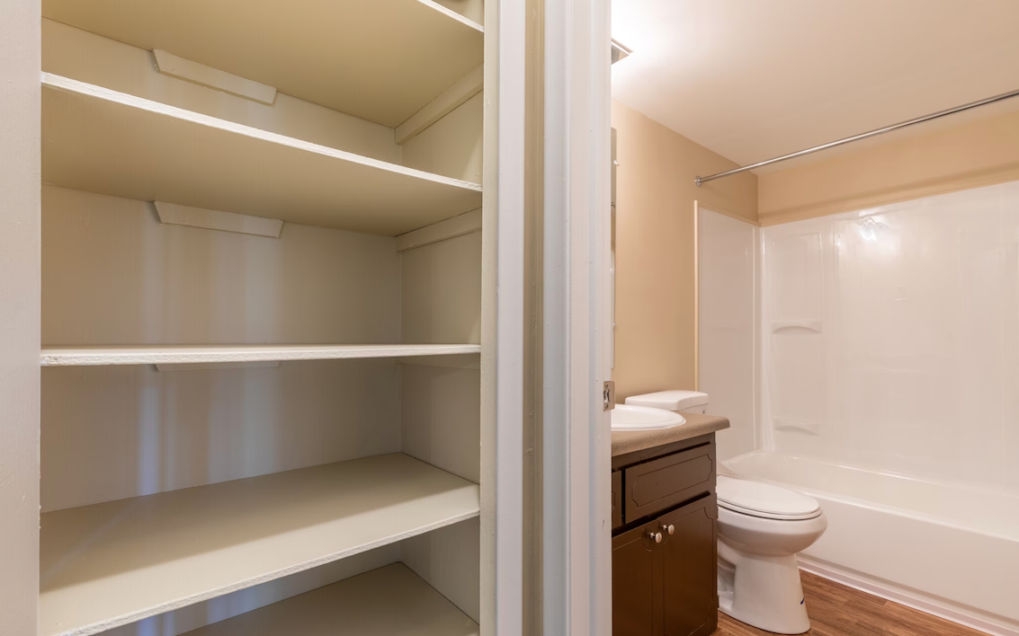Empty walk-in closet beside a beige bathroom with white shelves, a sink vanity, and a toilet.