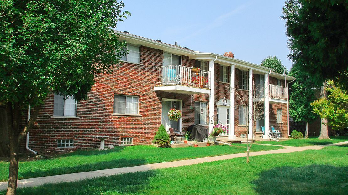 Brick apartment building with white columns and balconies, set in a green lawn with trees
