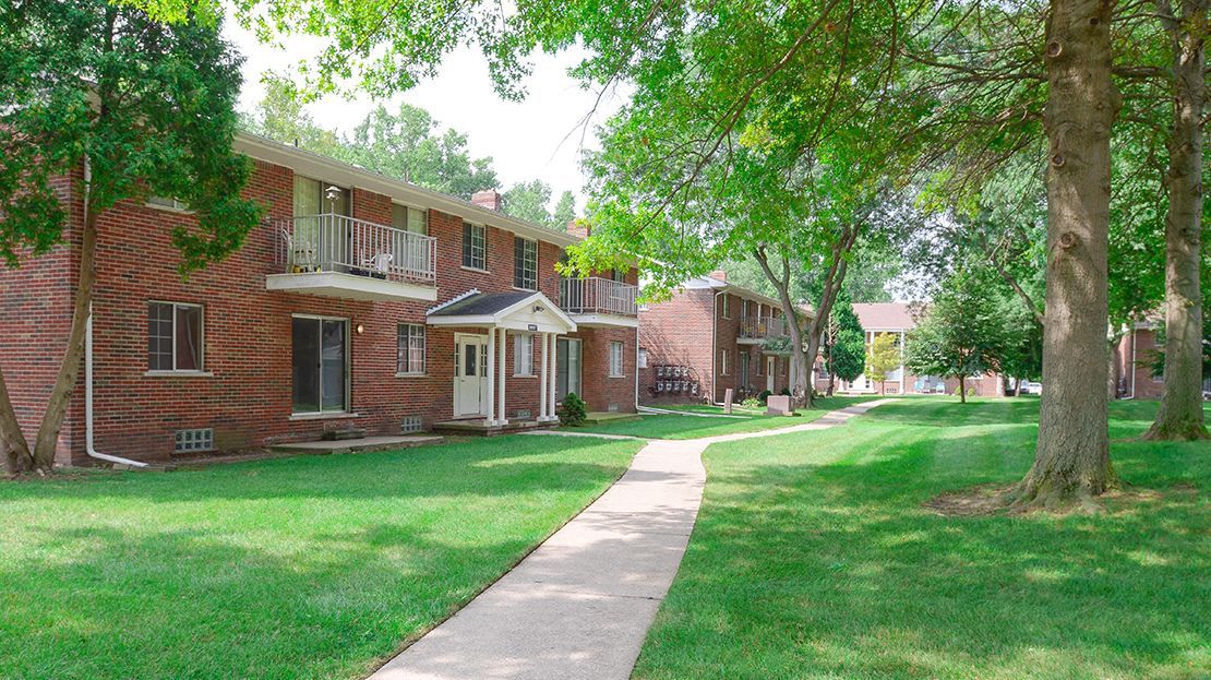 Brick apartment building with a sidewalk, green lawn, and trees in a sunny courtyard