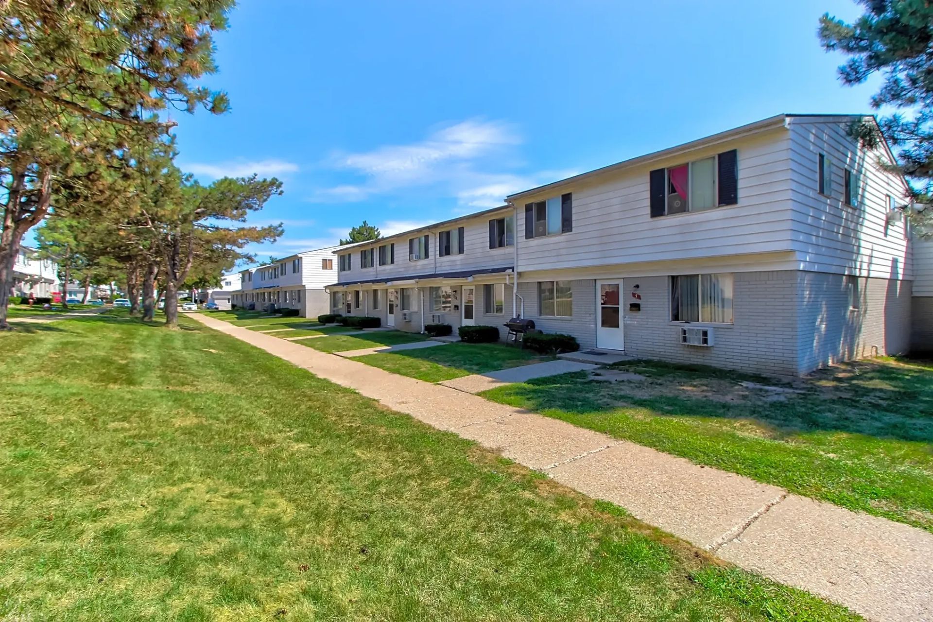 Apartment complex with beige two-story buildings, grassy lawn, and sidewalk under a blue sky
