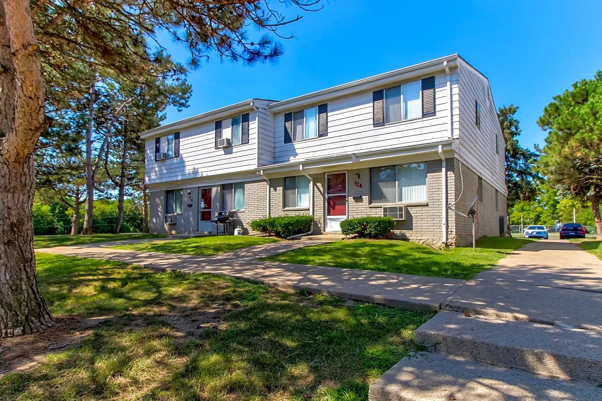 Two-story apartment building with white siding, trees, and a driveway on a sunny day