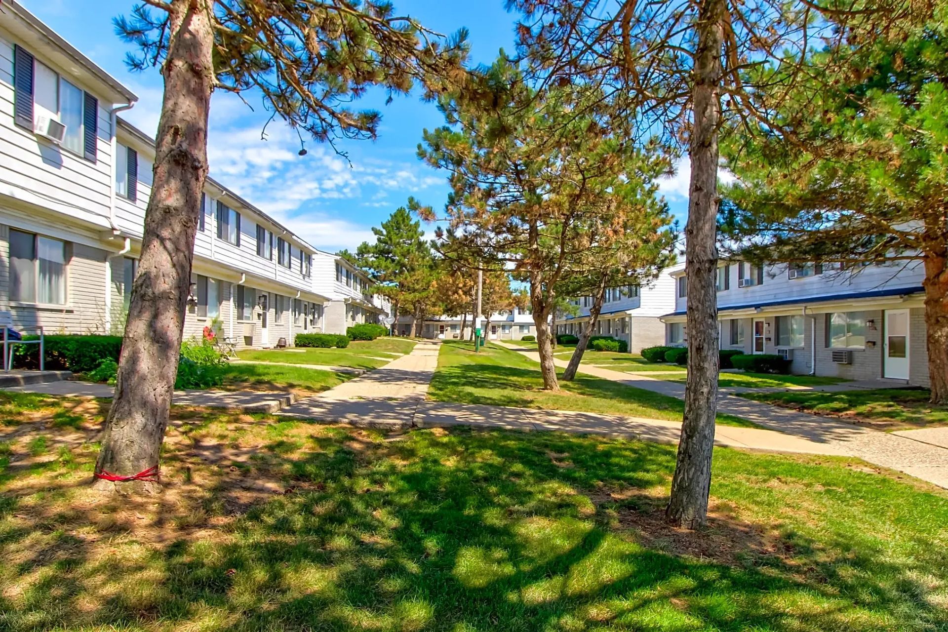 Apartment courtyard with trees, green grass, and walkways between white buildings under a blue sky