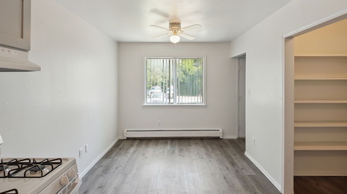 Empty white kitchen and living area with gray floors, a window, and a ceiling fan