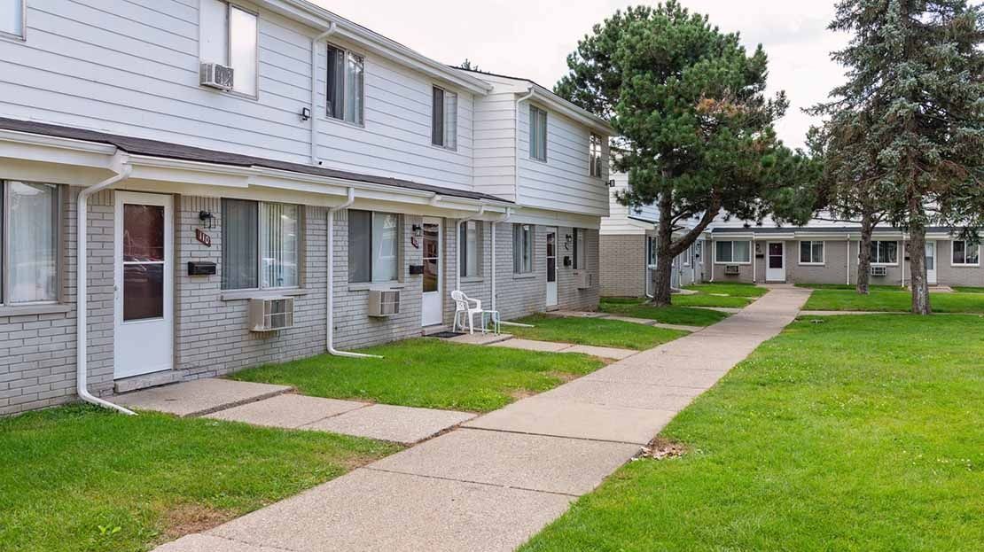 Row of light-gray apartment units with green lawn and a sidewalk in front
