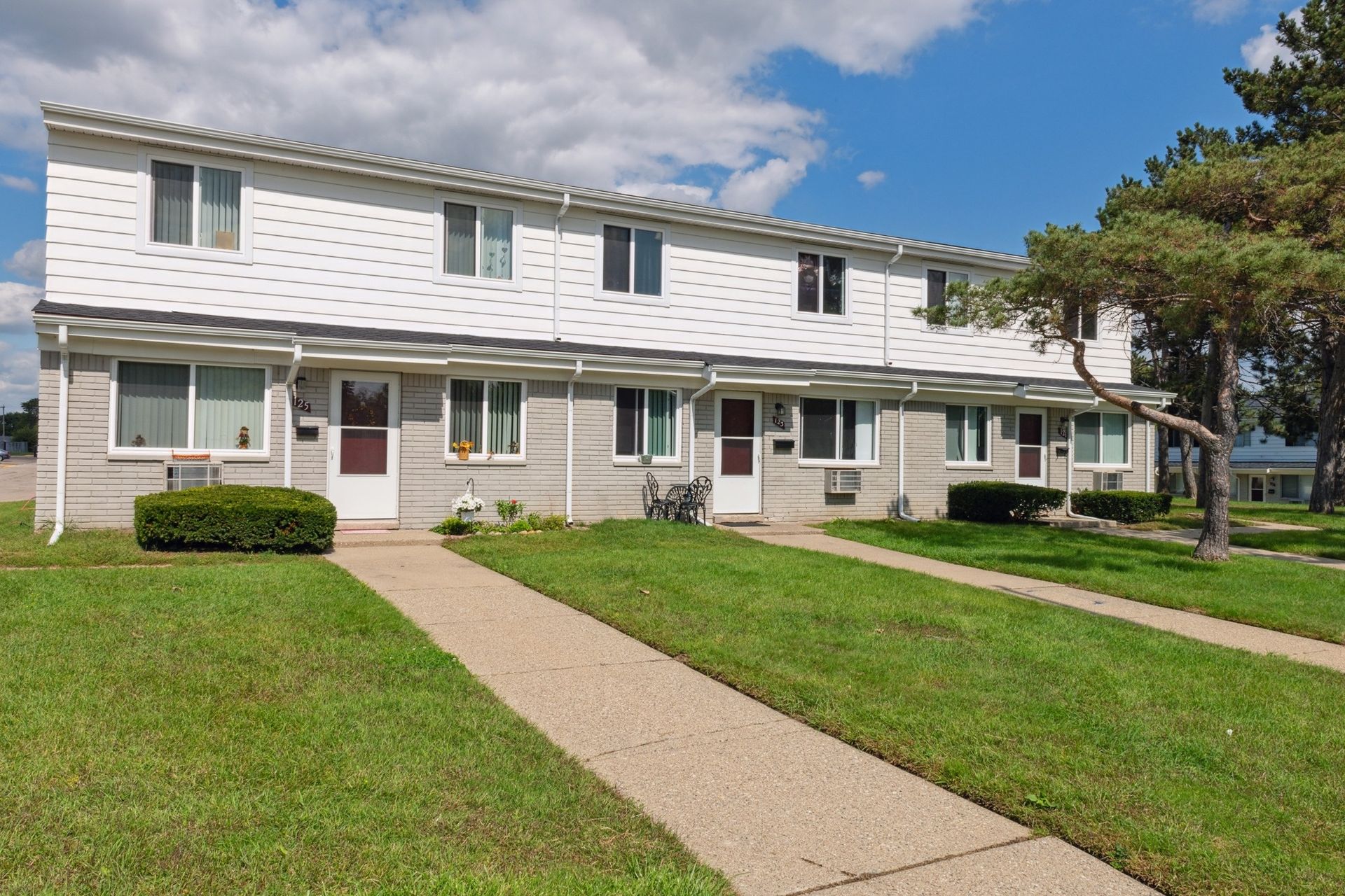 Row of two-story white apartment townhomes with green lawns and a sidewalk under a partly cloudy sky