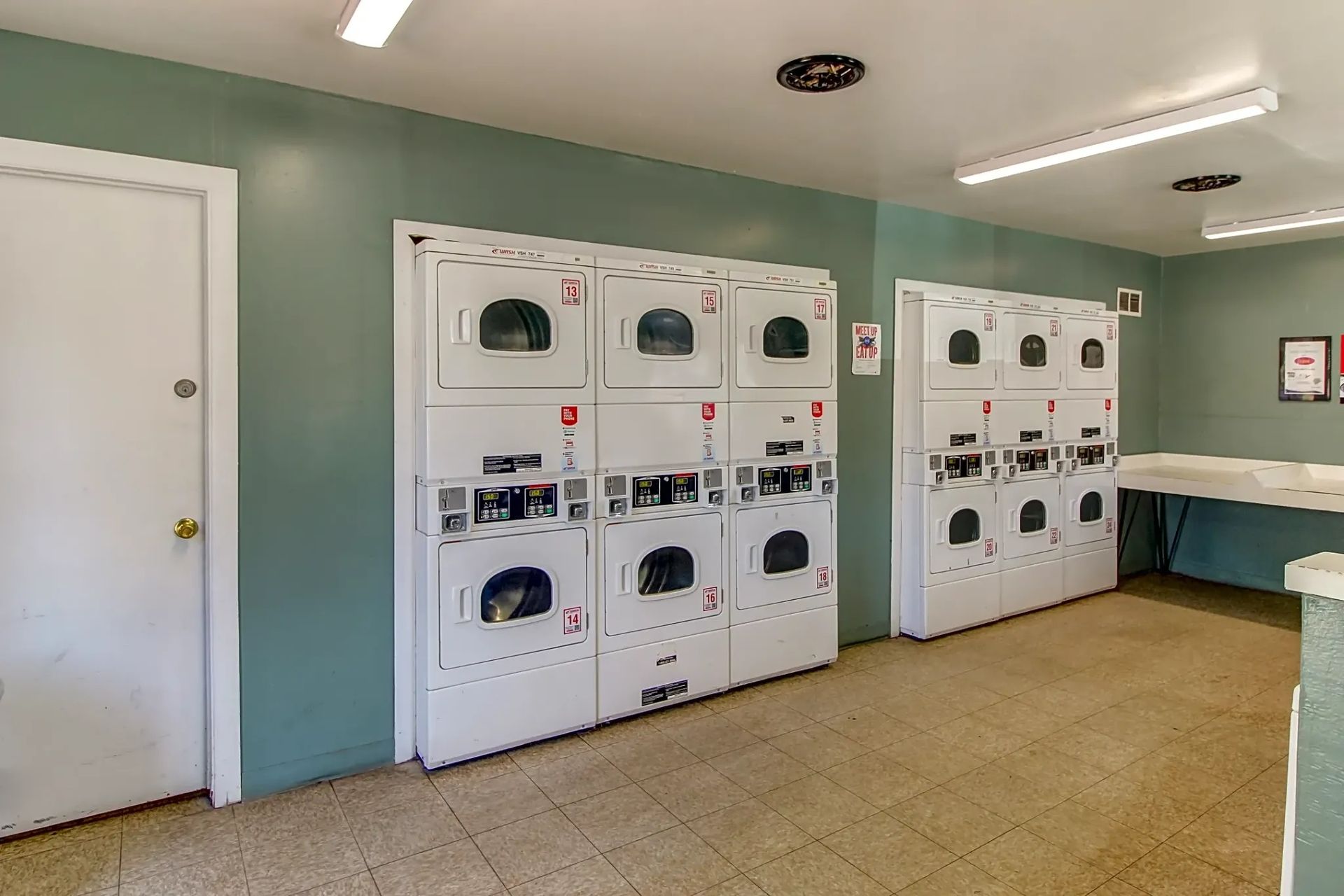 Laundry room with white dryers and washers against pale green walls, plus folding tables and a door.